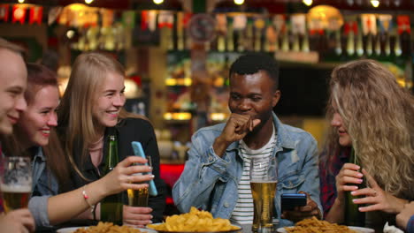 Das-Mädchen-Zeigt-Ihren-Freunden-An-Einem-Tisch-Sitzend-Den-Bildschirm-Eines-Smartphones-Und-Alle-Lachen,-Während-Sie-In-Einer-Bar-Sitzen.-Besprechen-Sie-Die-Neuesten-Gerüchte-Und-Klatsch,-Teilen-Sie-Fotos-Von-Reisen,-Zeigen-Sie-Lustige-Videos-Und-Fotos.